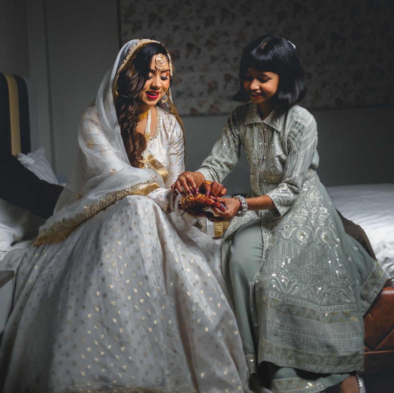 A bride in ivory laughs as a young girl adjusts her bangles on a bed
