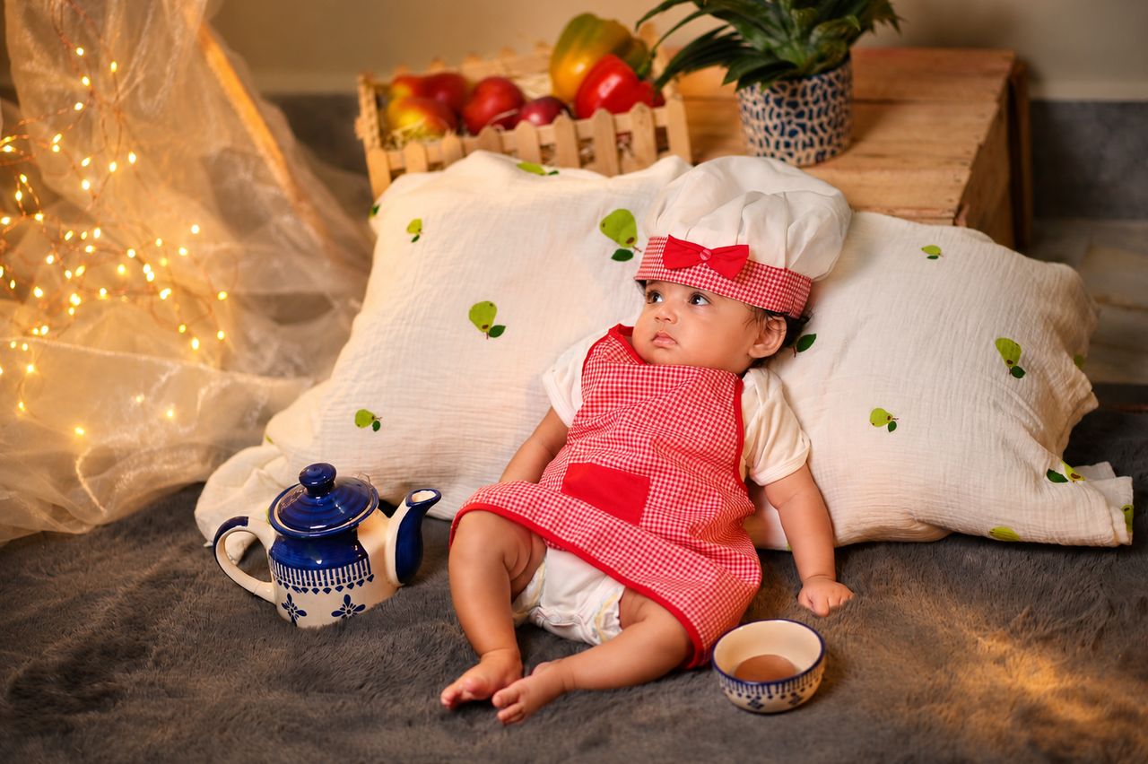 A baby girl in a little chef's apron and hat sitting among teacups and fruit