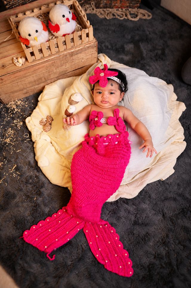 A baby in a pink crochet mermaid outfit lying on a cream blanket