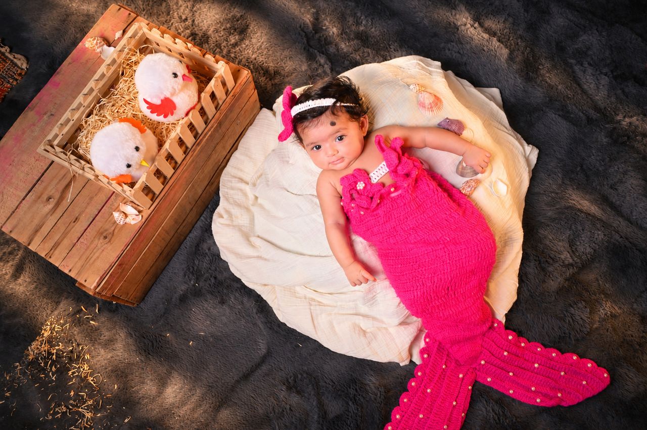 A baby in a pink mermaid outfit beside a wooden crate of soft toys