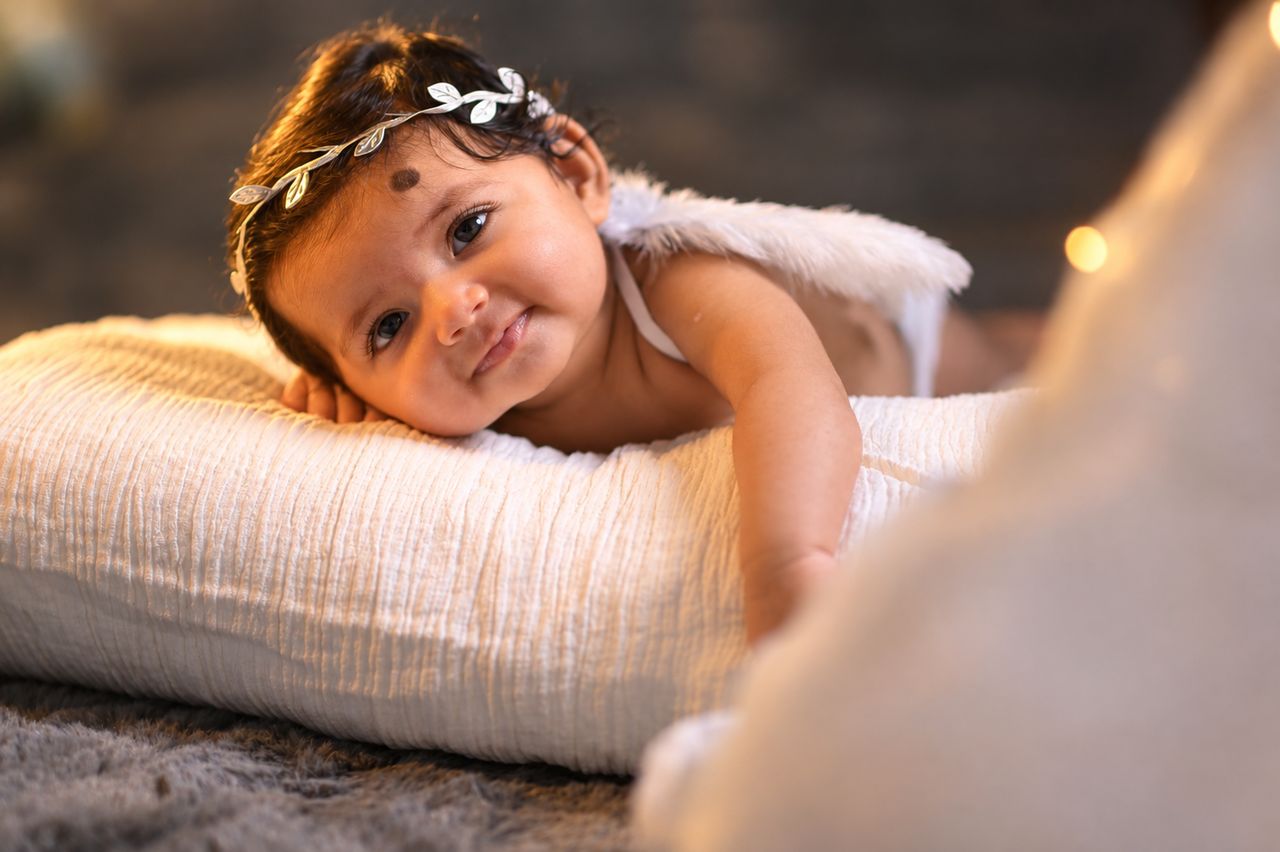 A smiling baby with a floral headband resting on a cream pillow