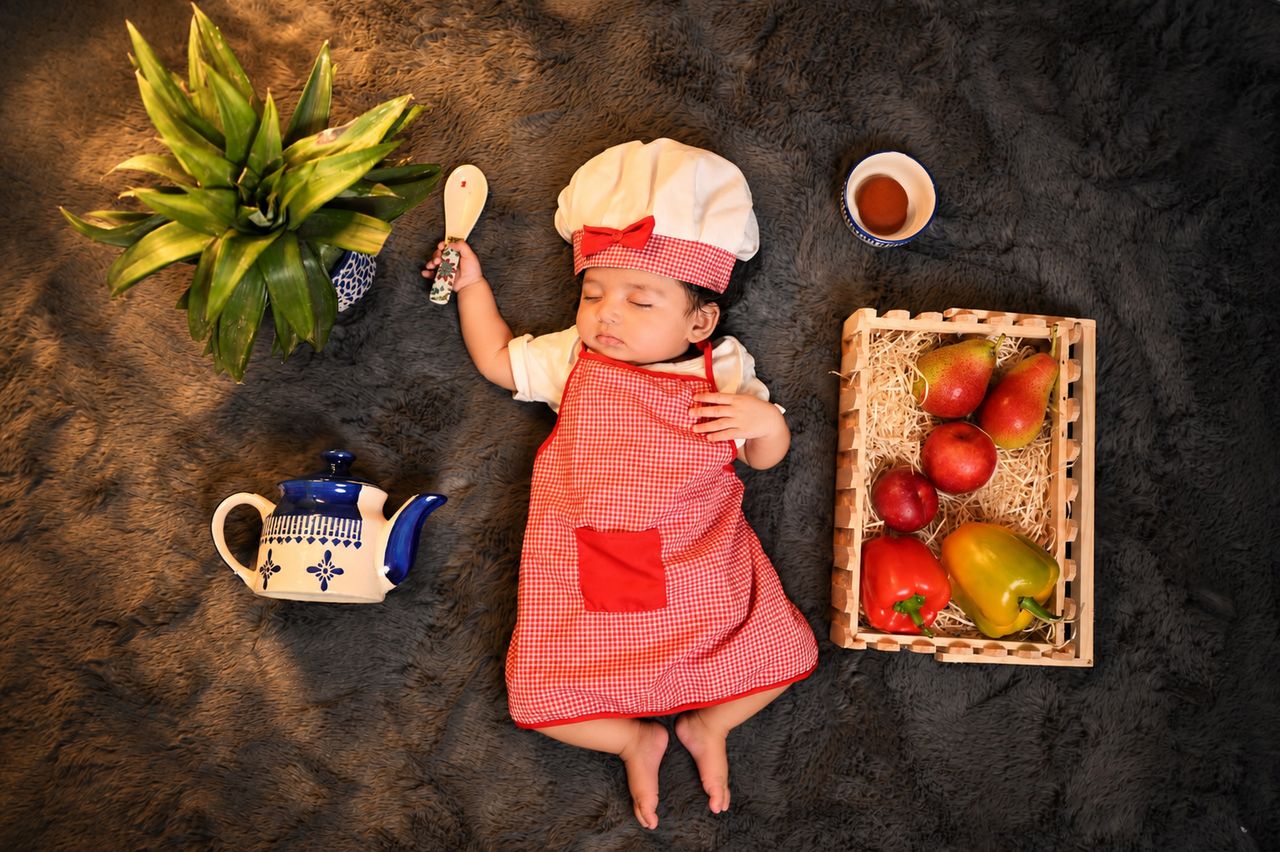 Overhead shot of a baby dressed as a little chef with a teapot, spoon and fruit basket