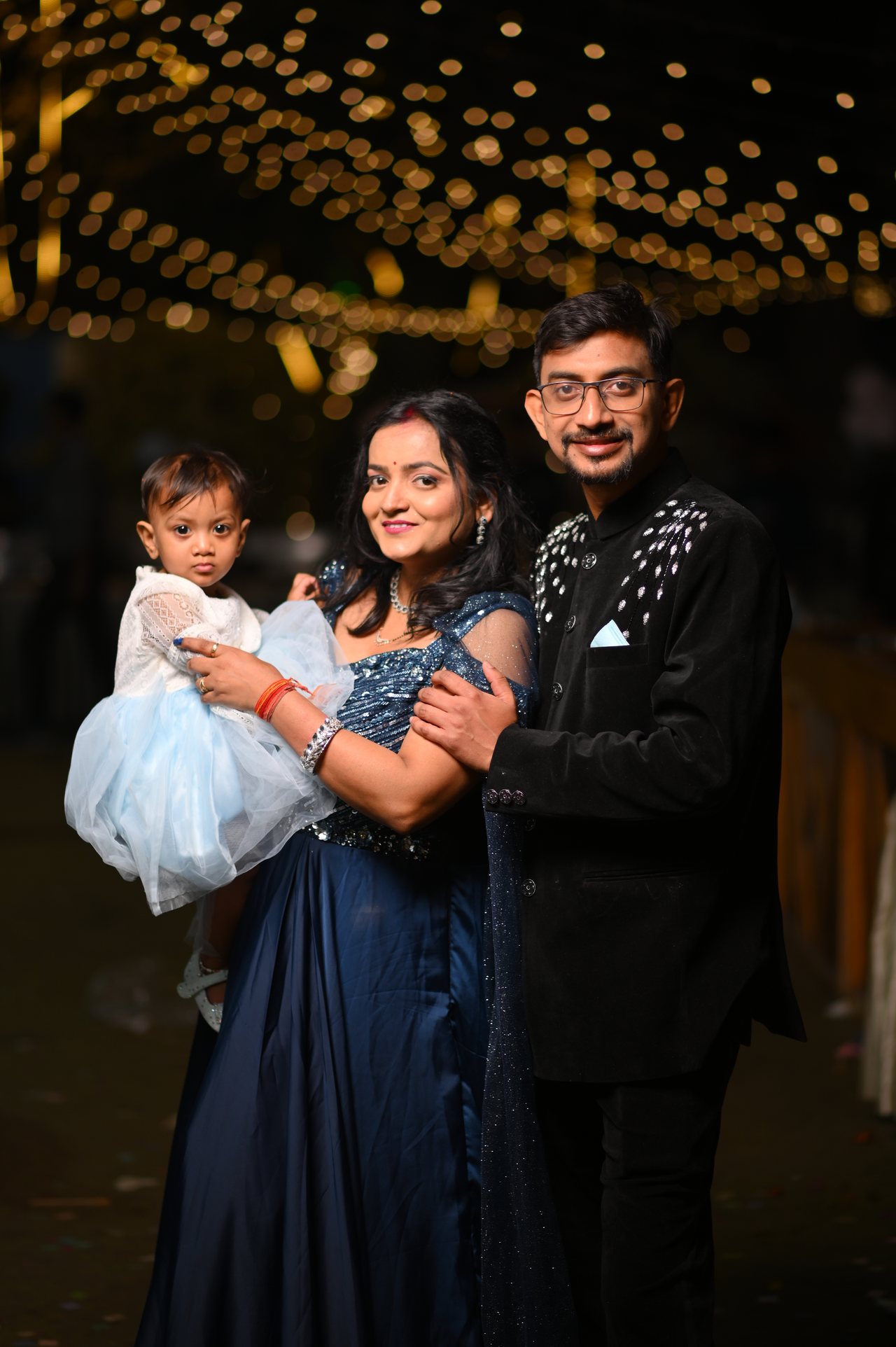 A mother and father hold their baby daughter under strings of warm lights