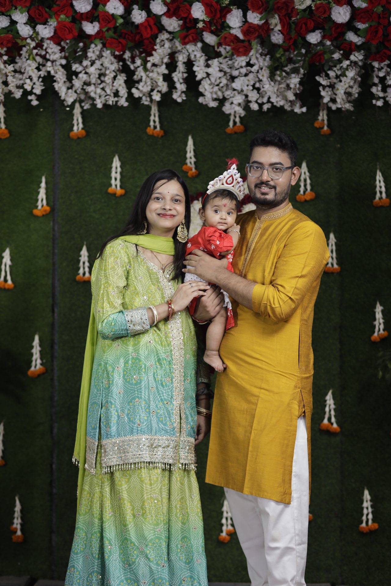 A couple hold their baby between them against a floral and grass backdrop