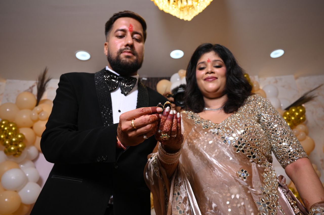 An engaged couple in a tuxedo and shimmering lehenga hold the rings between them