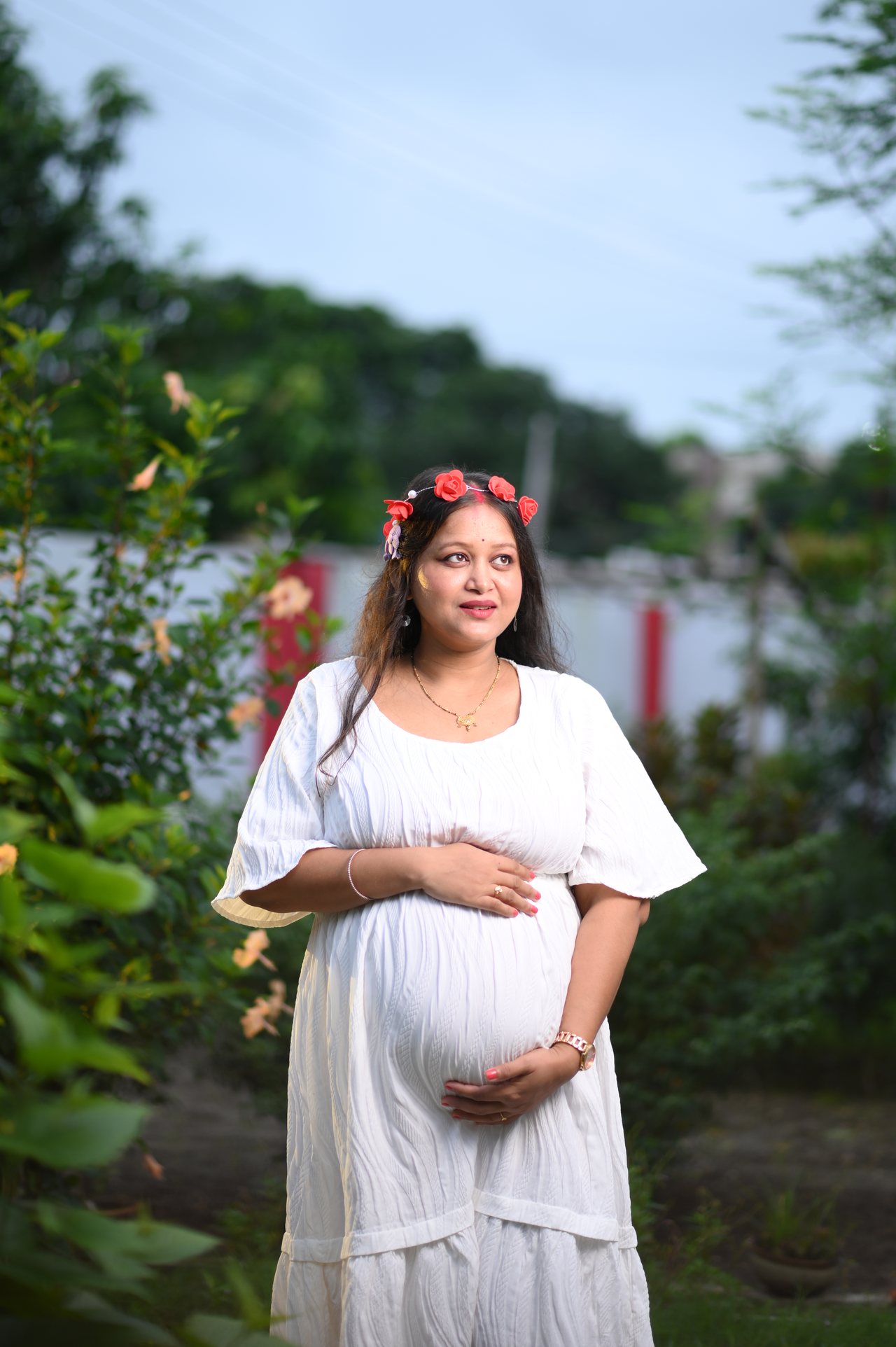 A mother-to-be in a white dress and flower crown cradles her bump in a garden