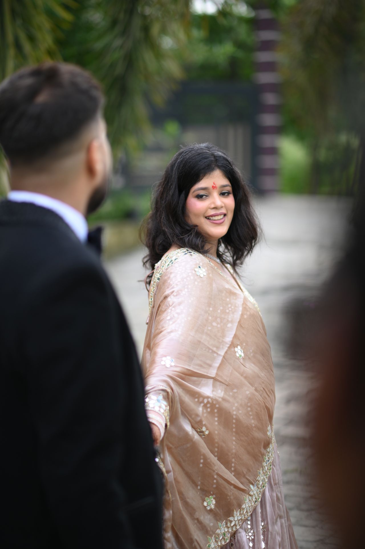 A woman in a champagne lehenga looks back at her partner in a garden