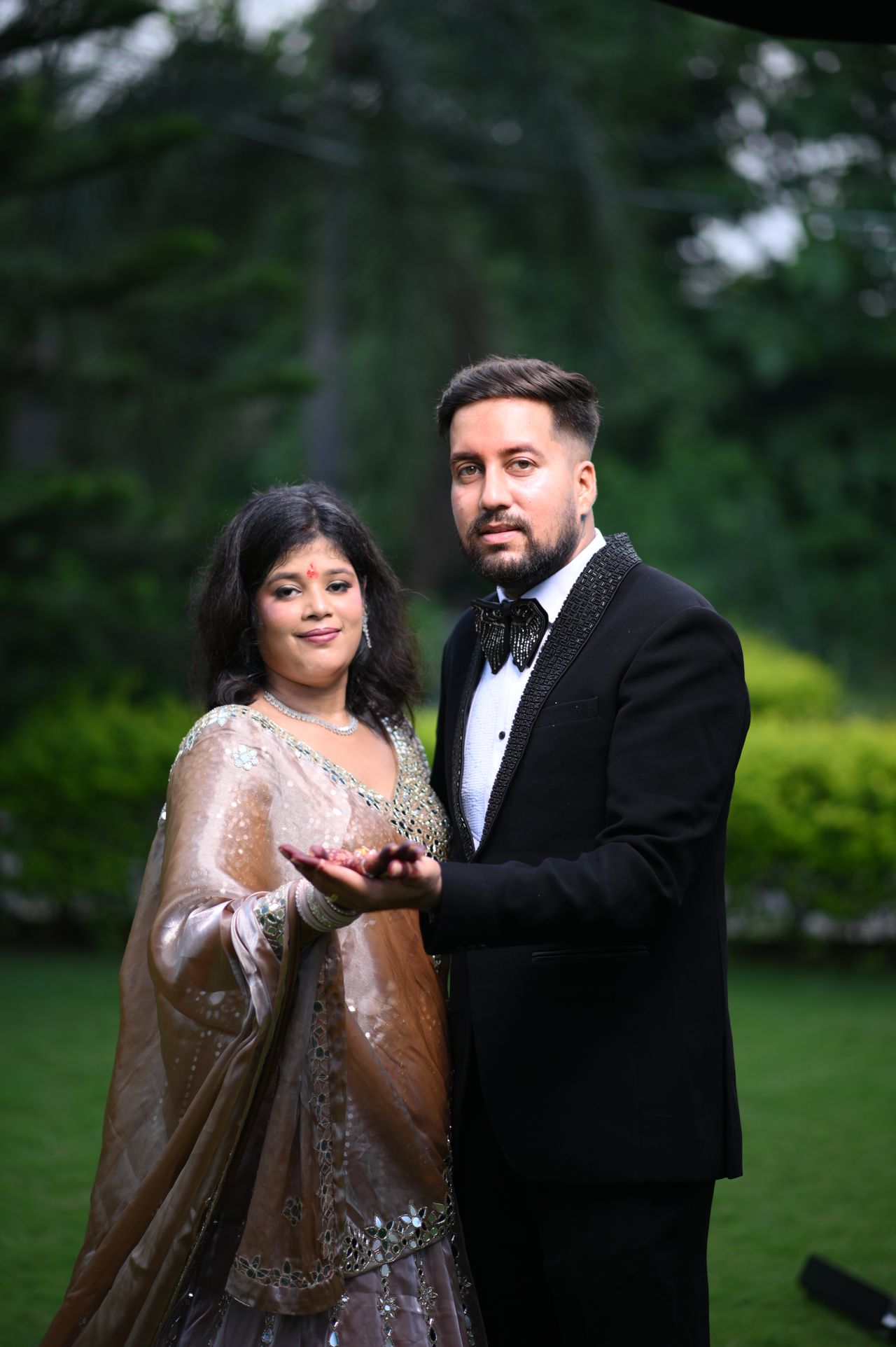 A couple stand together on a lawn, showing their rings