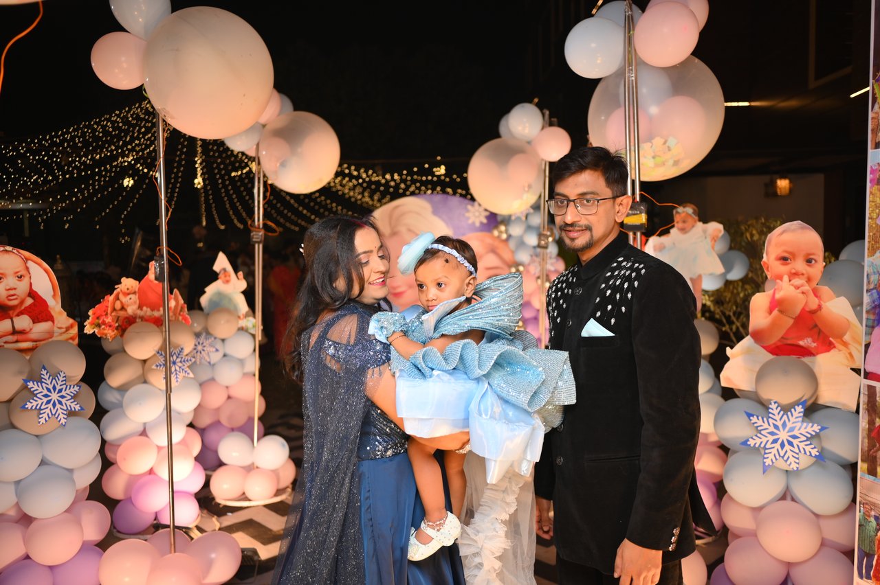 Parents hold their daughter in a blue dress in front of a pastel balloon backdrop