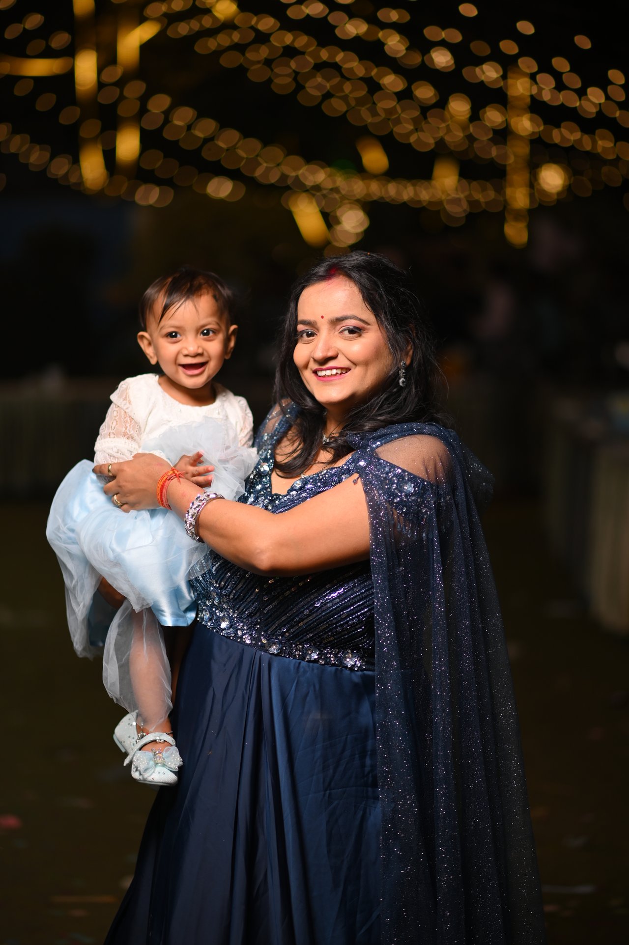 A mother holds her daughter beneath strings of warm lights
