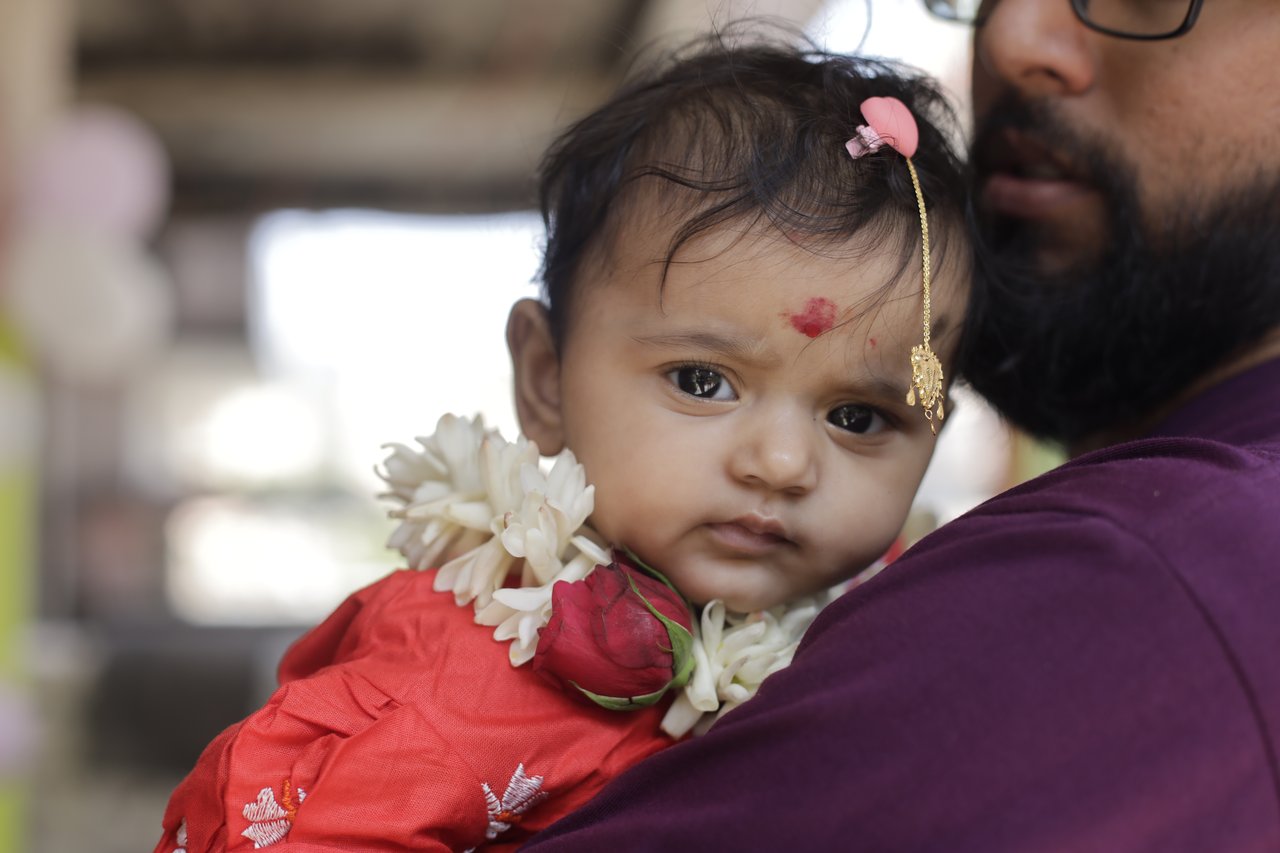 A baby in red with a floral garland rests on her father's shoulder