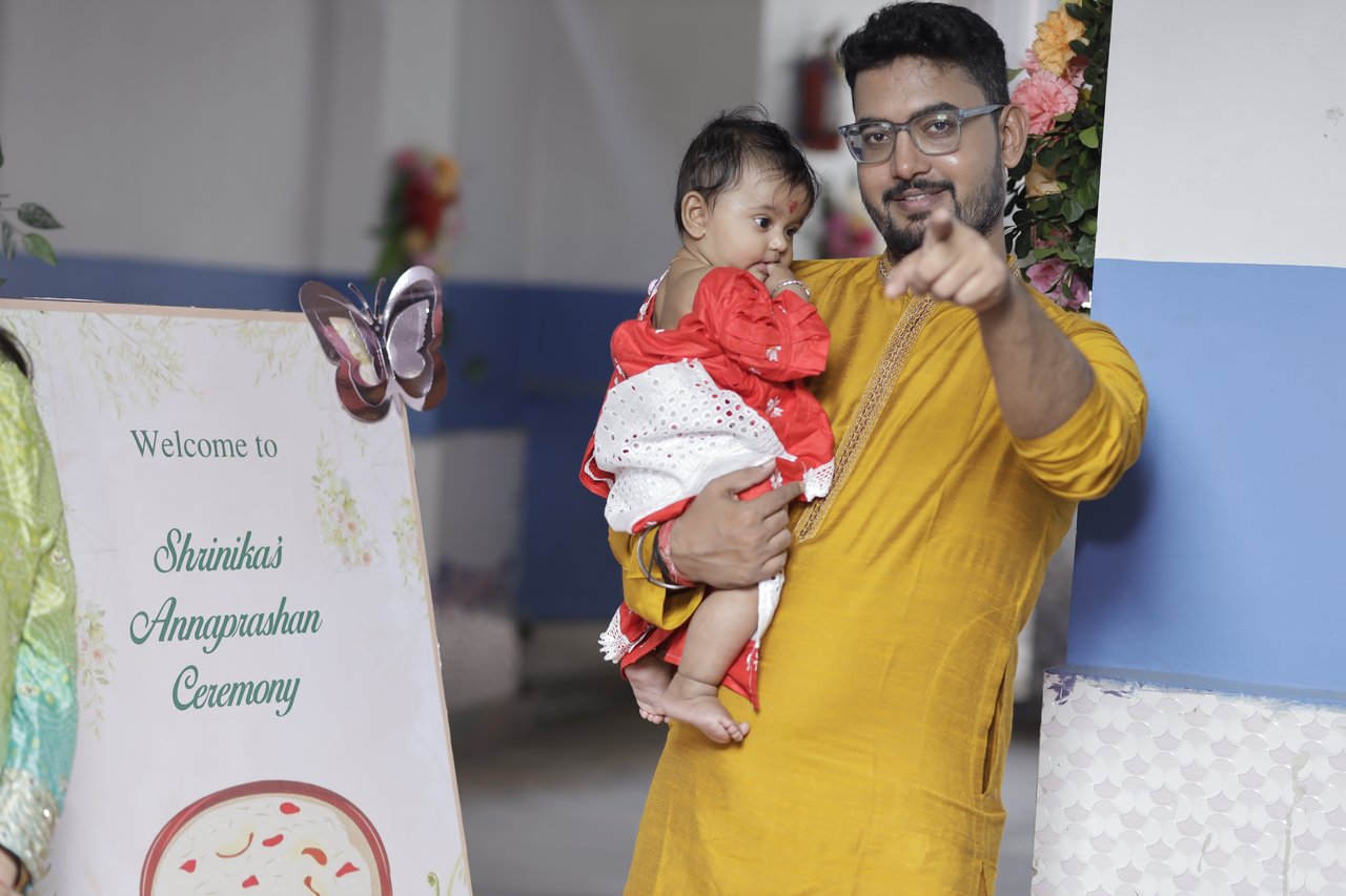 A father points at the camera, holding his baby beside the welcome board