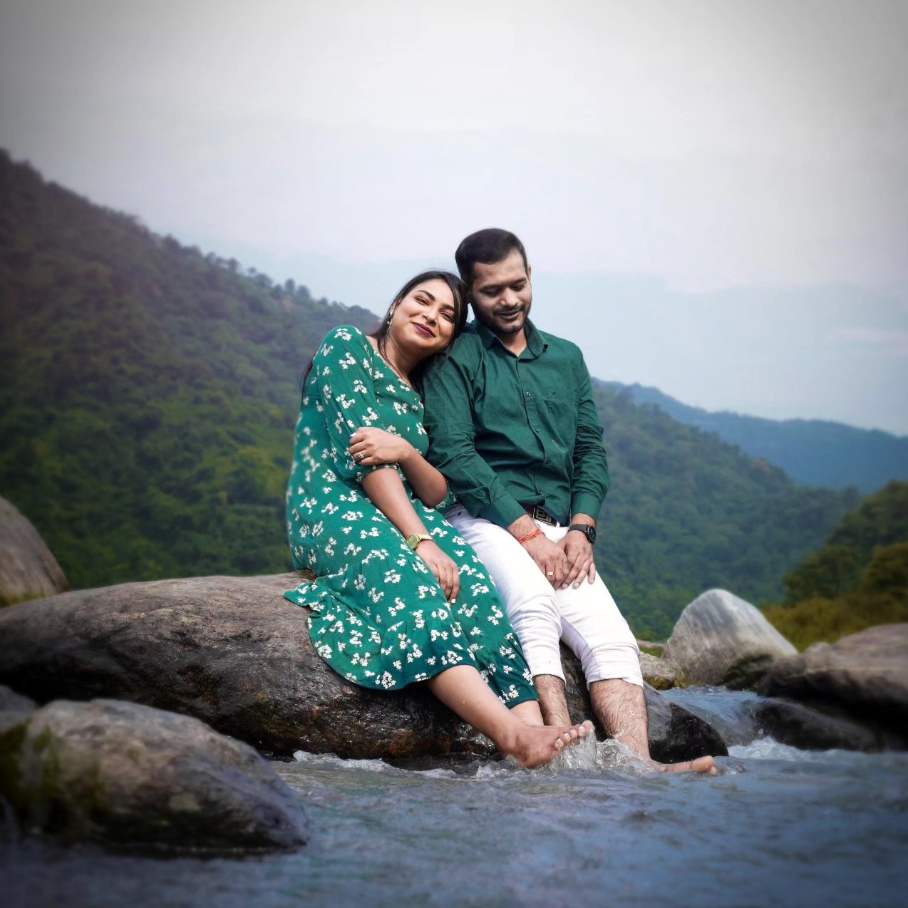 A couple sit on rocks by a river with green mountains behind