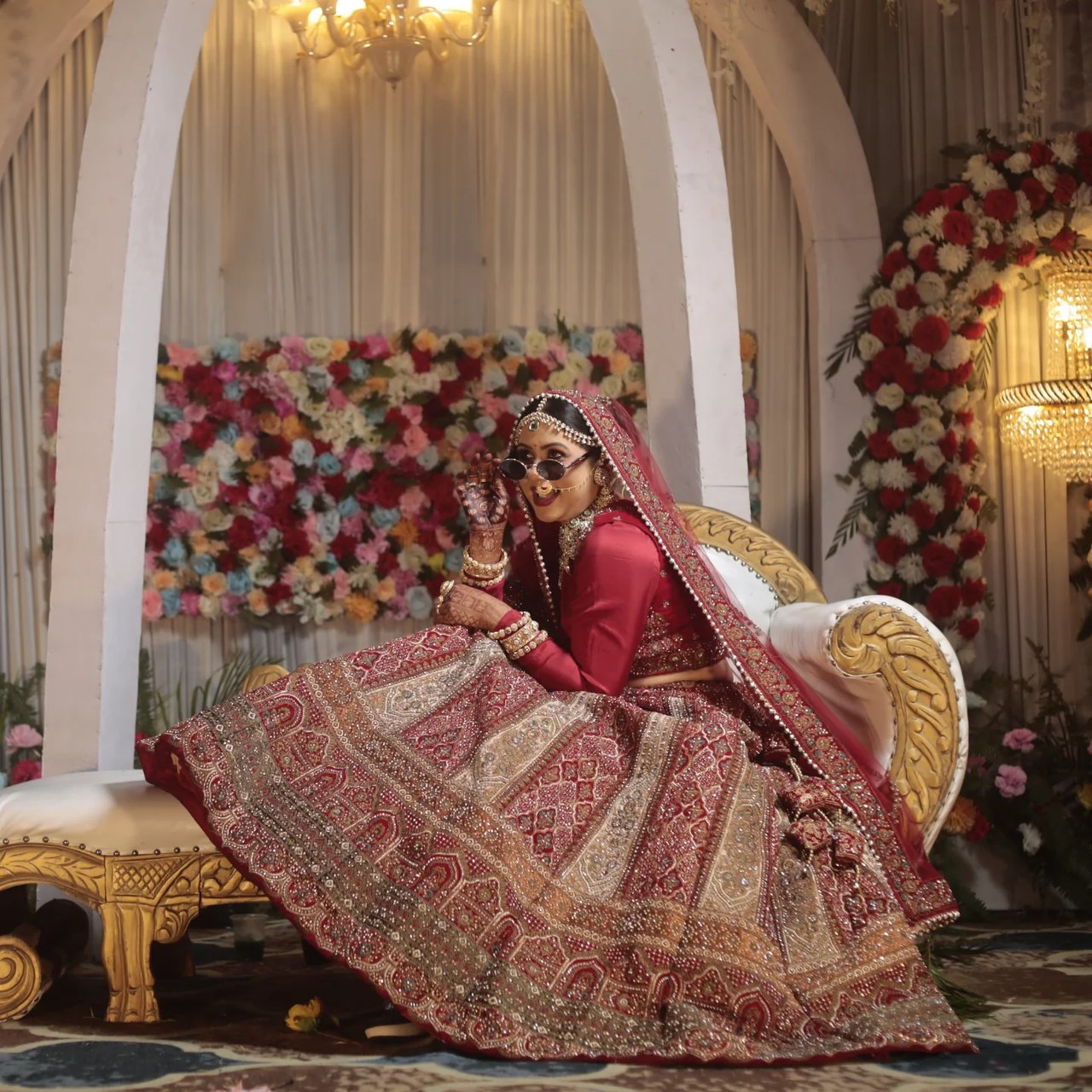 A bride seated on a decorated stage sofa in a red lehenga
