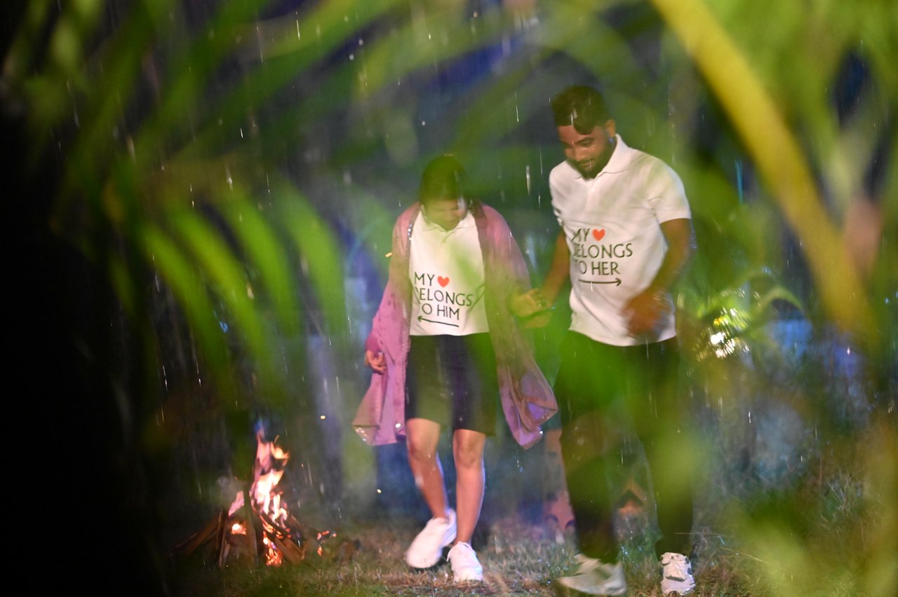 A couple walk past a small fire among tall plants at dusk