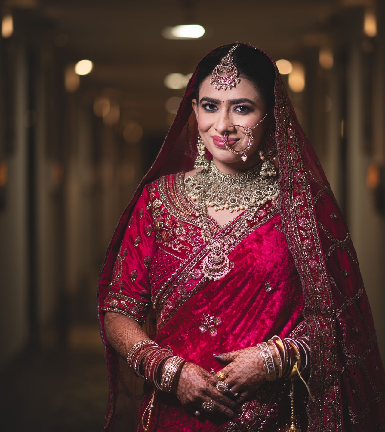 Bride in a crimson lehenga and heavy jewellery, softly lit in a dark corridor