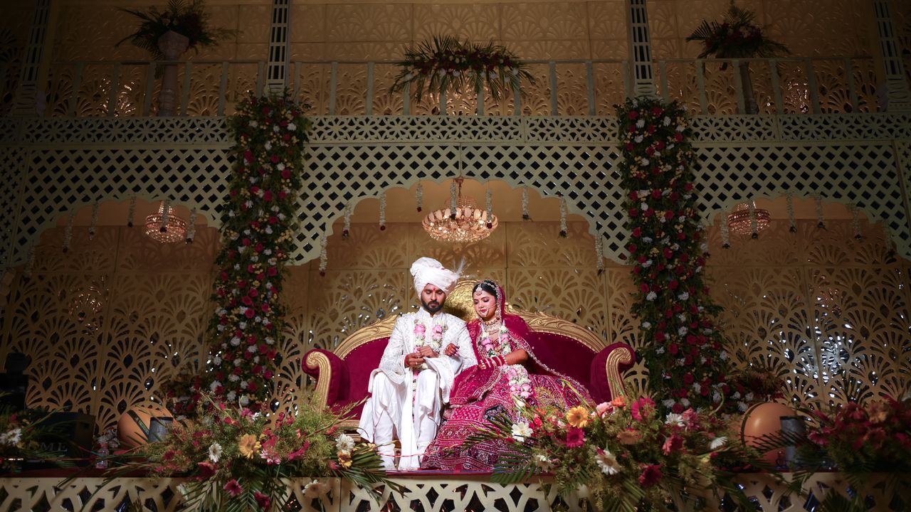 Bride and groom seated on a red sofa beneath an ornate floral stage
