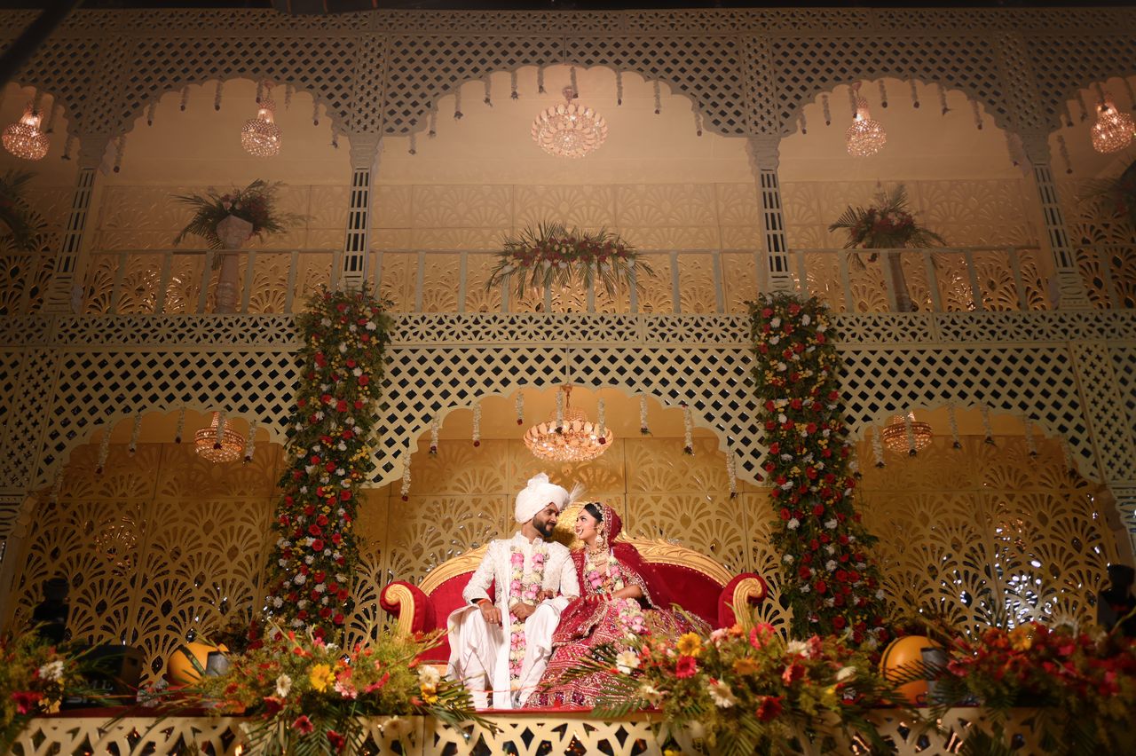 Bride and groom seated on an ornate reception stage of carved arches, chandeliers and flowers