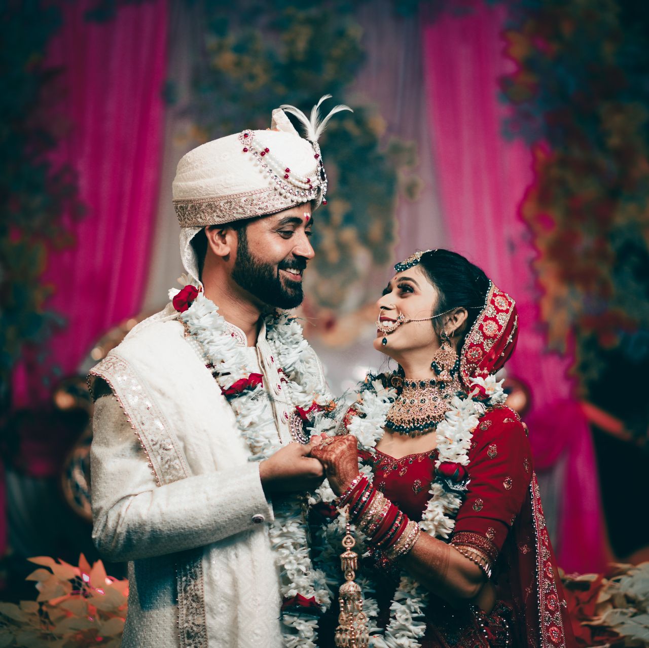Bride and groom in garlands holding hands and smiling at each other under a pink floral mandap
