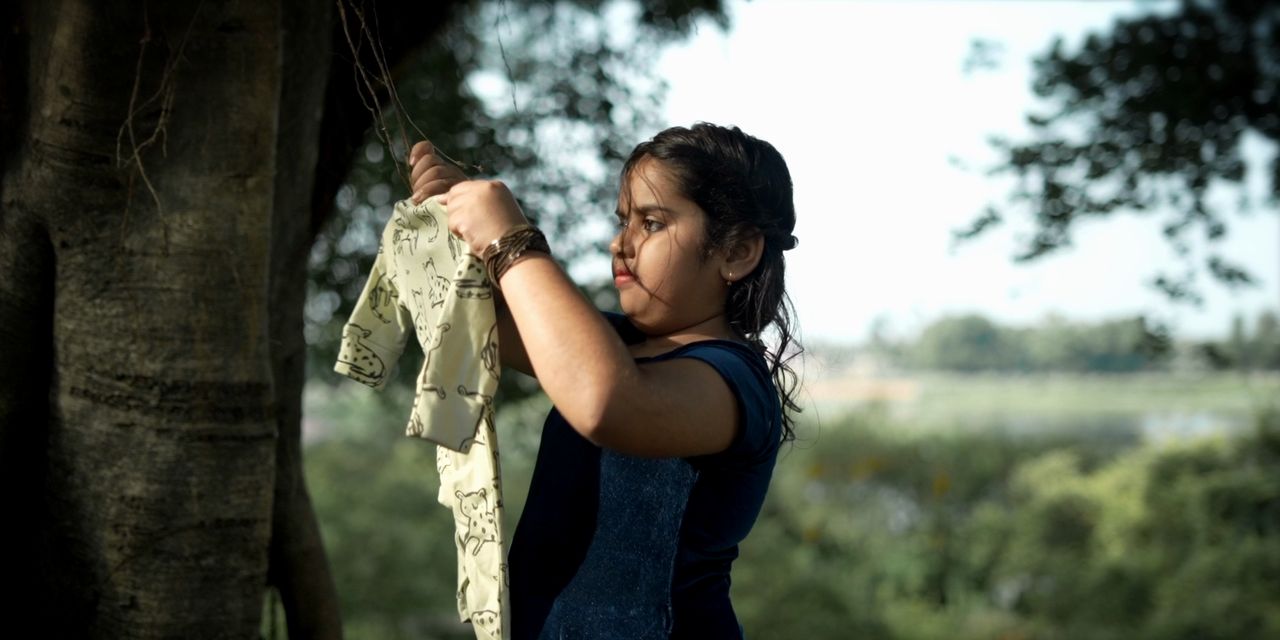 A young girl hangs tiny baby clothes on a line beneath a tree