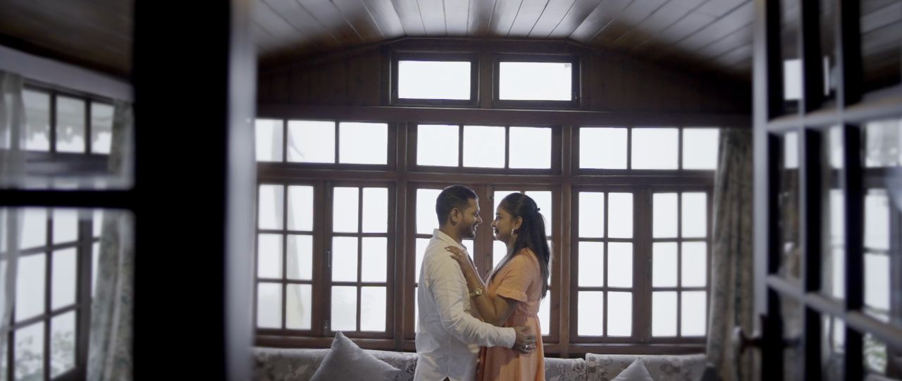 A couple embrace in front of tall wooden windows in a hill-station cottage