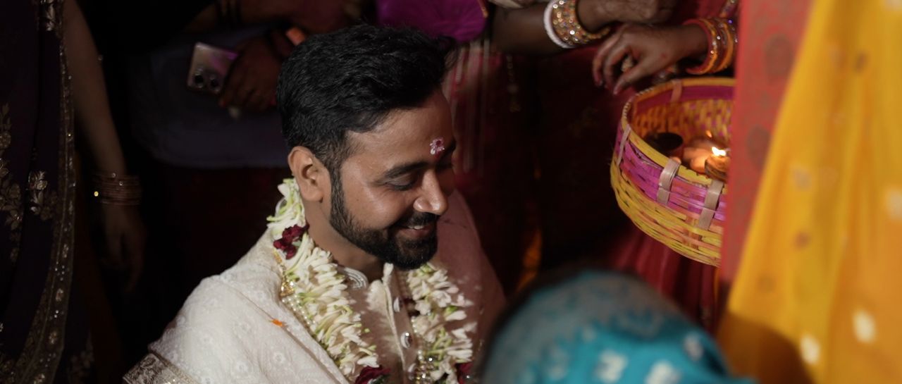 The groom smiles during a wedding ritual, garlands around his neck
